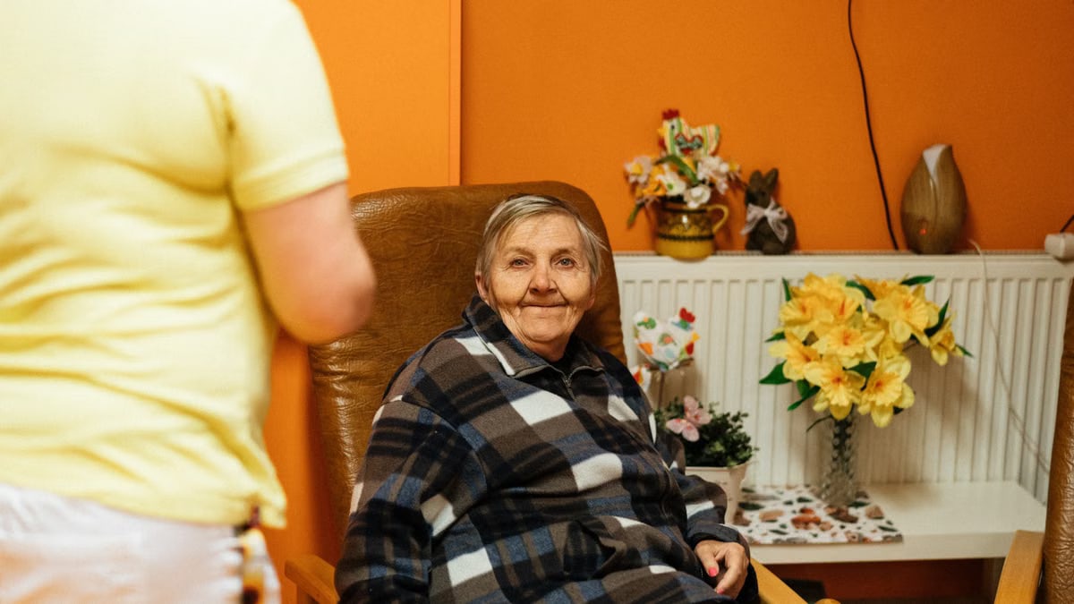A warm scene of a senior woman sitting in an armchair at a retirement home with a caretaker nearby.